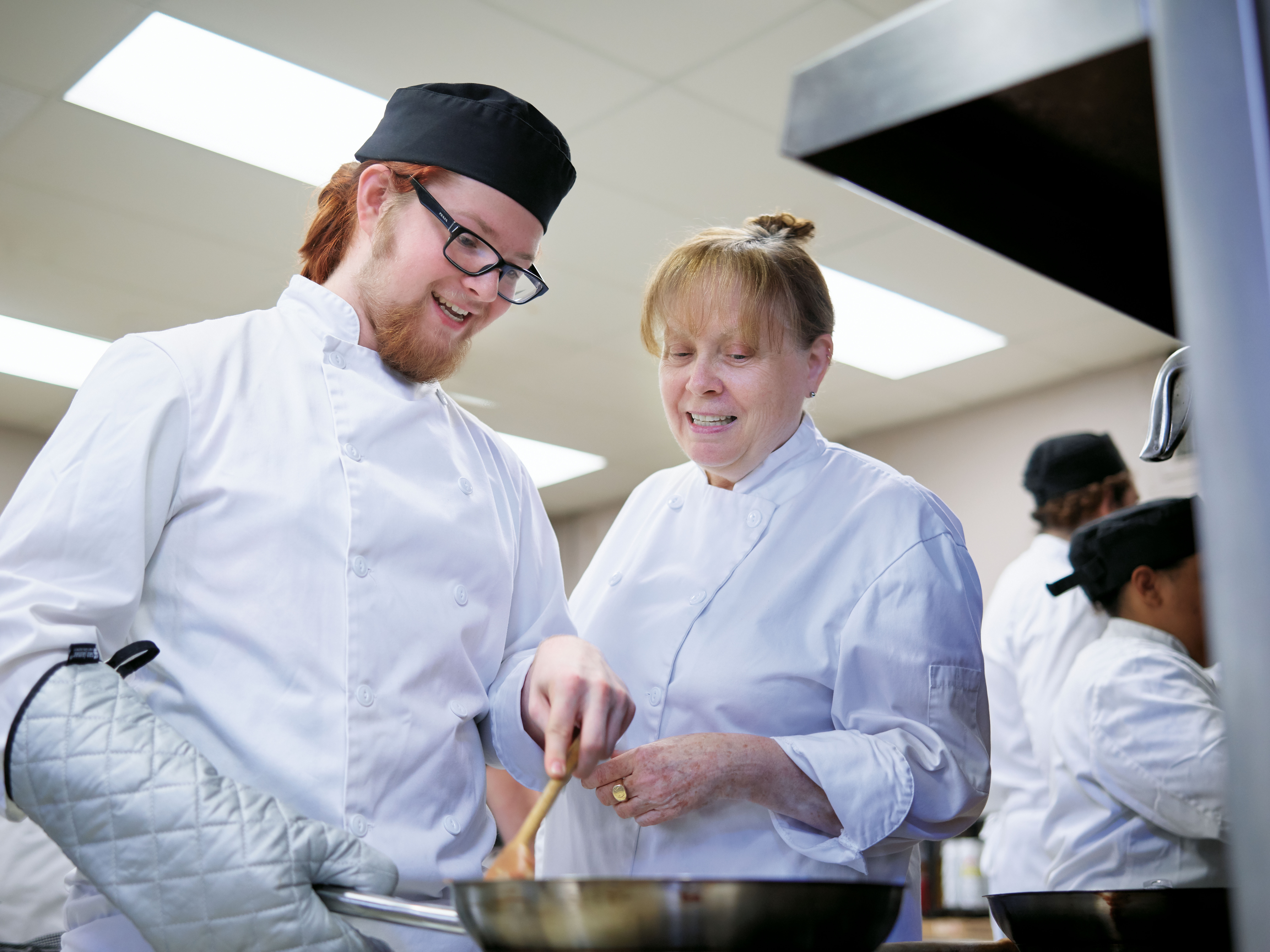 Photo of two adults dressed in chef attire. One is using a spoon to stir contents in a saucepan.