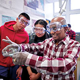 Photo of a teacher and two students in a classroom wearing safety goggles. The teacher has gloves on to pour a substance from a bottle.