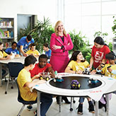 Photo of Josée Landriault, OCT, smiling in a suit with arms crossed. Josée stands in between two tables in a classroom with students sitting and standing.