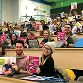 Photo of a large group of teacher candidates in a lecture hall, smiling and holding brochures and bags.